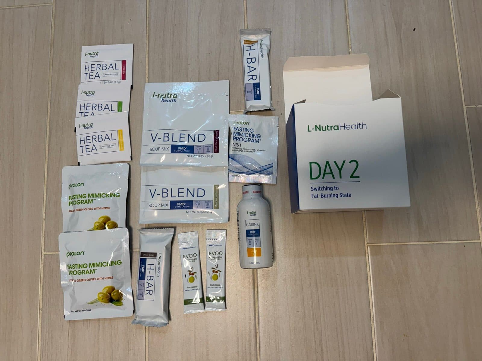 The day 2 Prolon kit laid out on a floor: herbal teas, two soup mixes, olives, two nut bars, olive oil, a drink, a supplement packet, and the Day 2 box.
